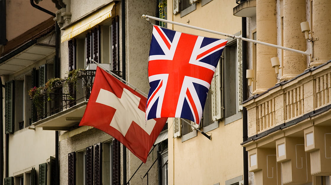 British and Swiss flags fly next to eachother from an upper floor in a old-fashioned apartment building with balconies and window shutters.