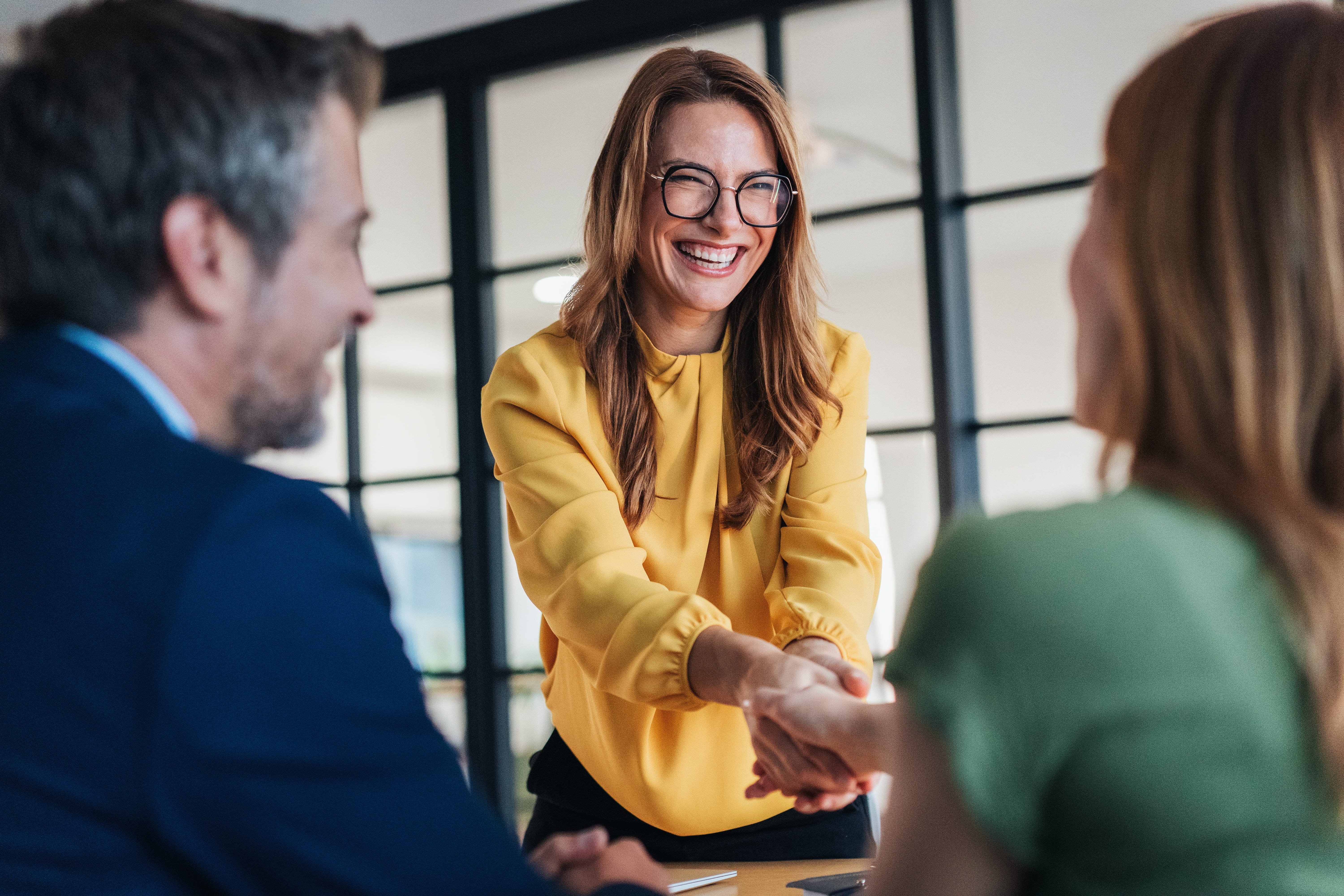 A woman wearing a yellow blouse and glasses smiles and shakes another woman's hand. 