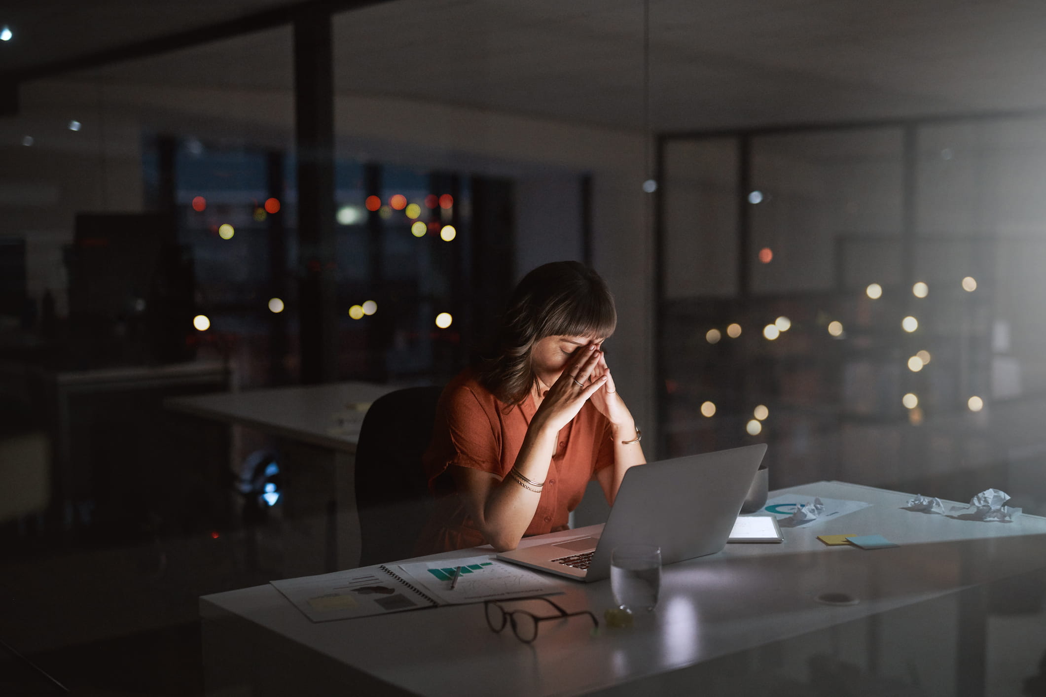 A young woman sits with her hands to her face, alone in a darkened office.