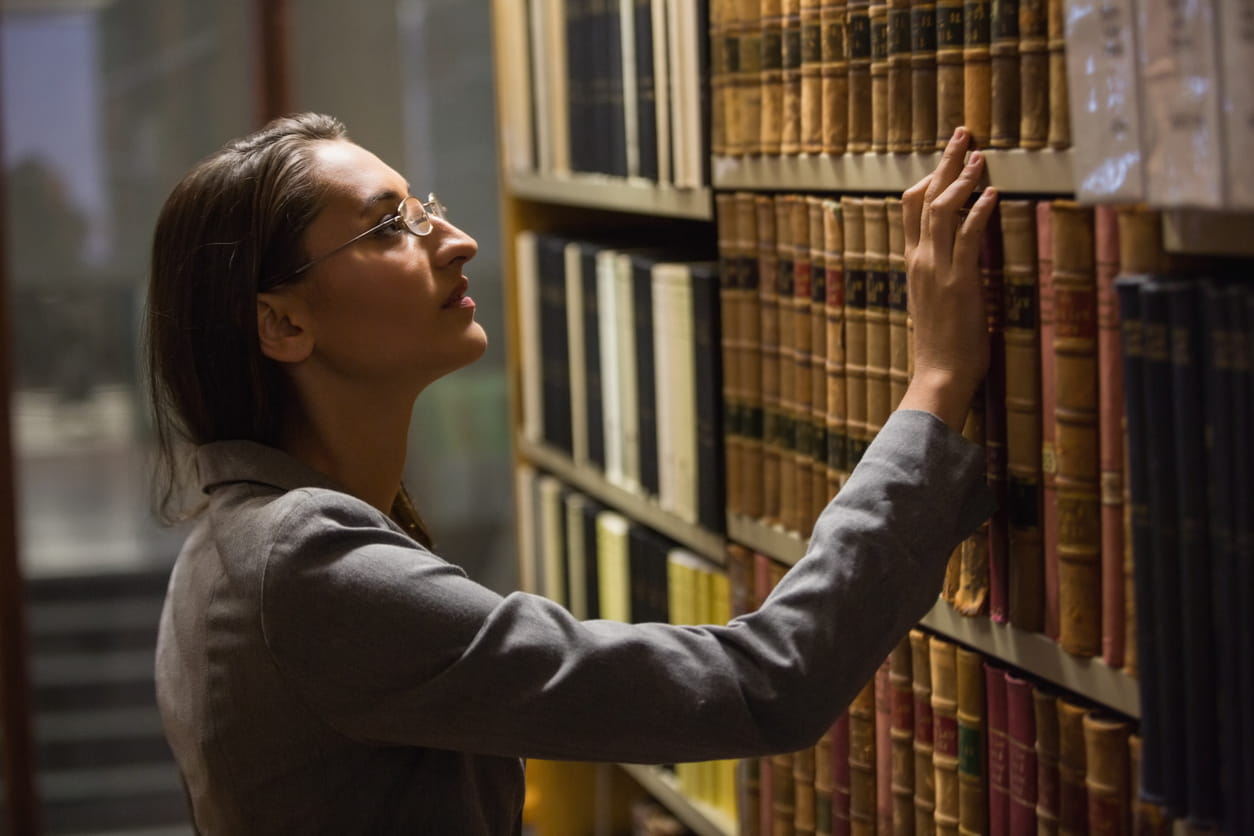 A woman in a grey suit and glasses picks out a book at the library.