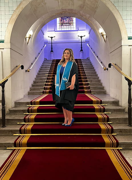 Solicitor Olivia Rogers wearing graduation robes stands on a grand staircase in the Law Society