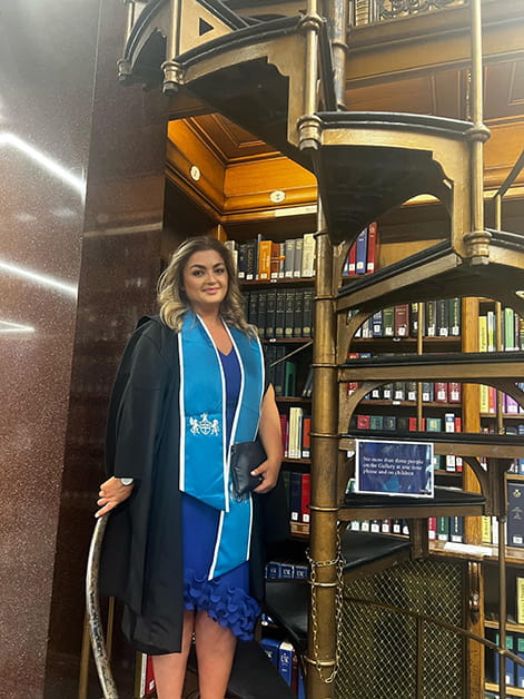 Solicitor Olivia Shaw wearing graduation robes stands on a spiral staircase in the Law Society library