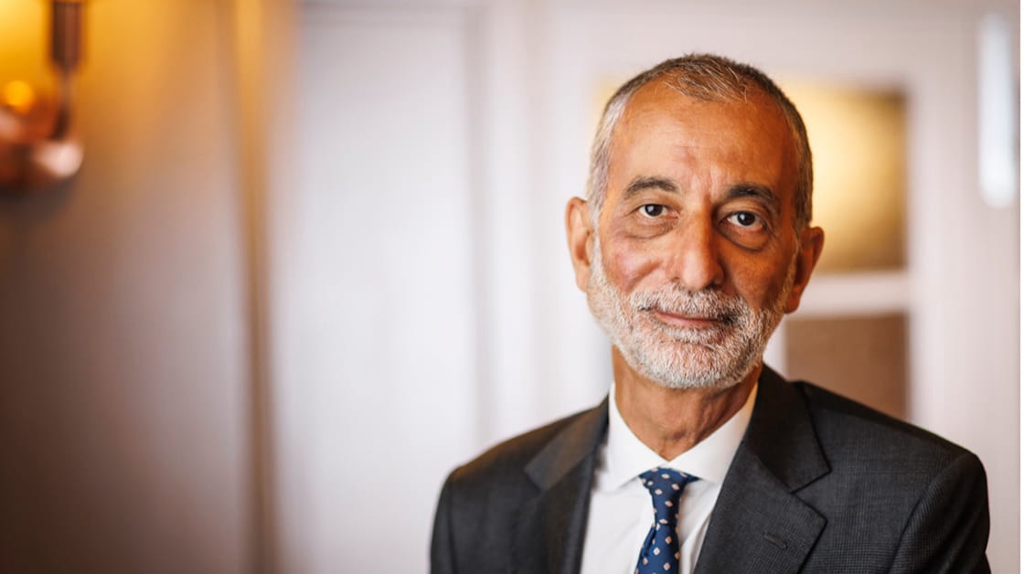 Imran Khan is a South Asian man with grey hair and a grey beard. He is wearing a black suit with a white shirt and blue patterned tie. He is smiling with his mouth closed. 