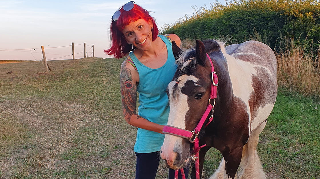 Arwen is a white woman with bright red hair. She is standing in a field with her horse wearing a blue vest top and sun glasses on her head. She has tattoos on her right arm and is smiling. 