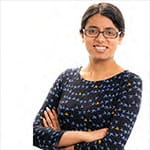 Coralie is wearing a black and white dress, she is wearing glasses and is smiling with her arms crossed.