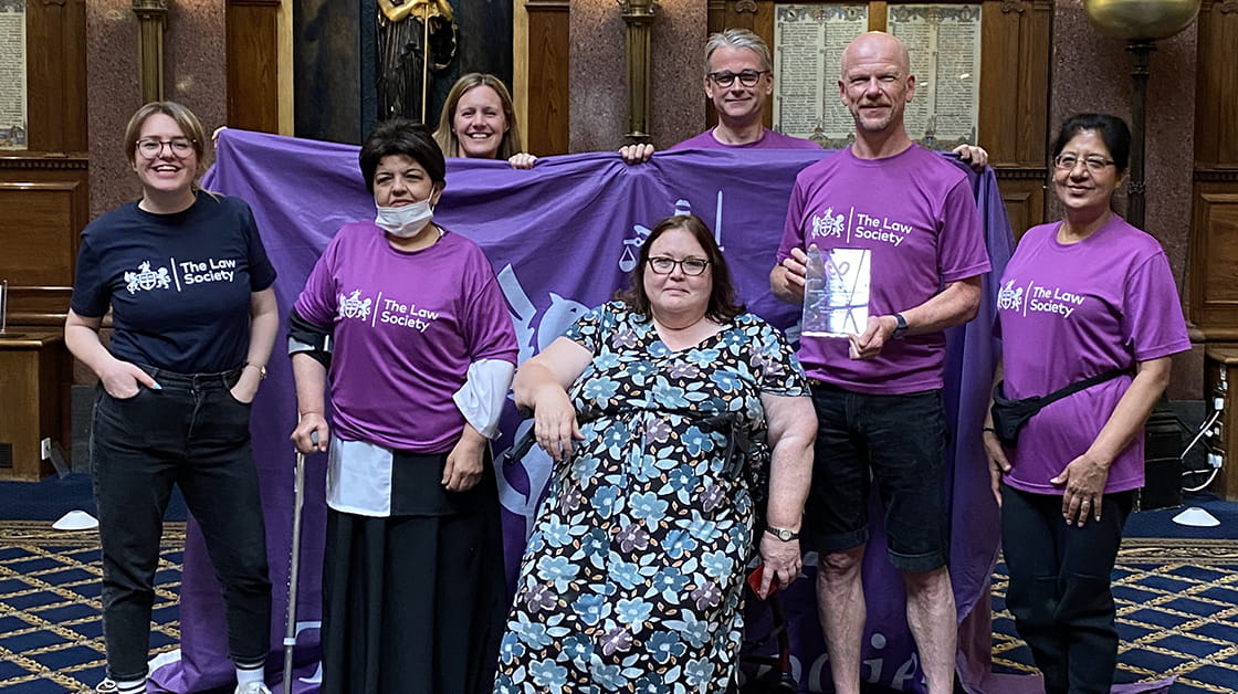 A diverse group of people wearing purple tops with the Law Society Logo written in white on the front. A man and a woman are holding up a purple banner, another man is standing in front holding a glass trophy.