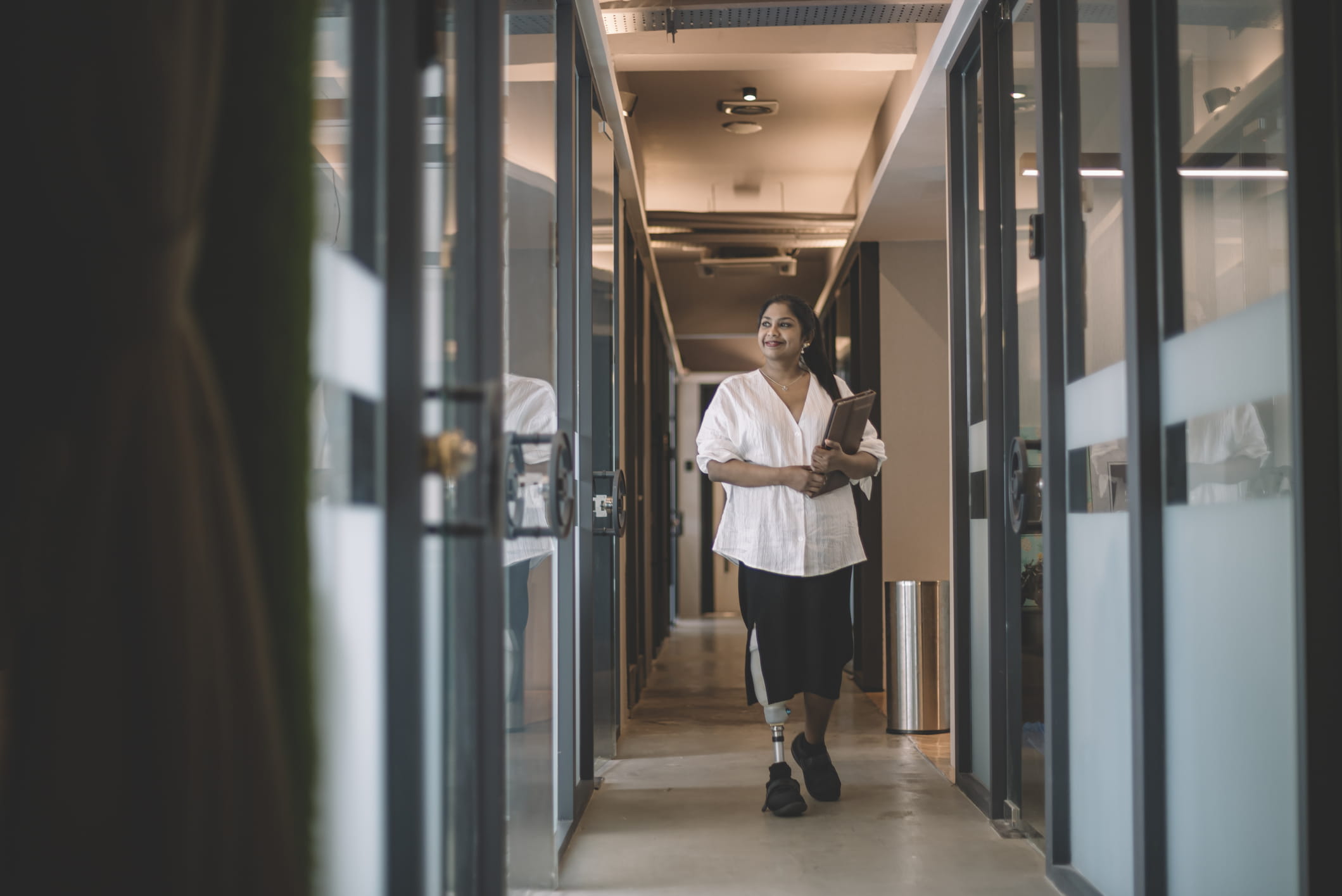 An Asian woman with a prosthetic leg is walking through a hallway in an office building. She is wearing a white shirt, a black skirt and black shoes. She is smiling and carrying an electronic tablet.