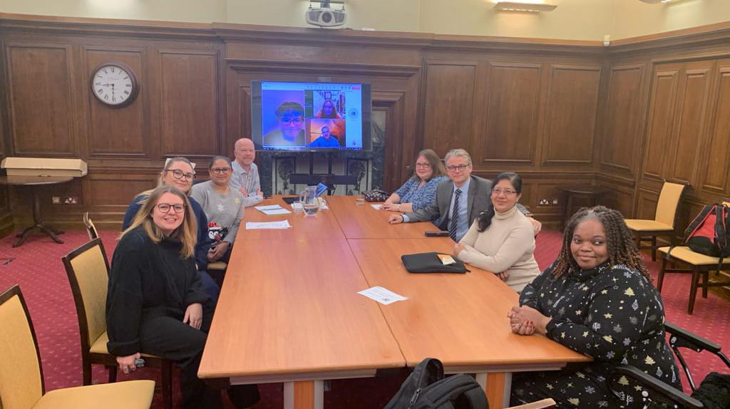 A diverse group of people are sitting around a big table. Theres a screen behind them with people on a teams call. The room is wood panelled with a red carpet. 