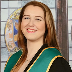 Hannah Clifford has blonde hair and is wearing a graduation robe, with a stained glass window in the background.