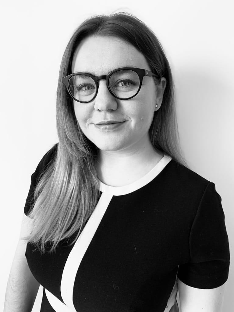 Lizzie Hardy is a white woman with long fair hair. She is wearing a black and white dress and glasses. She is smiling with a closed mouth. 