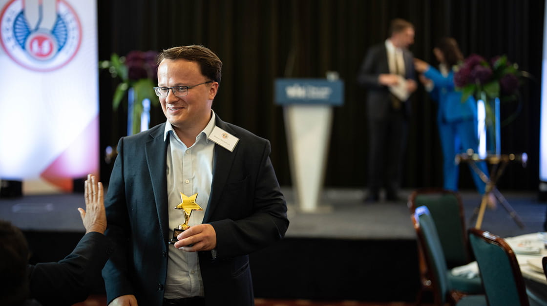 Martin is a white man, with brown hair and glasses. He is wearing a navy blue suit and a light blue shirt. He is walking and smiling, holding a trophy.