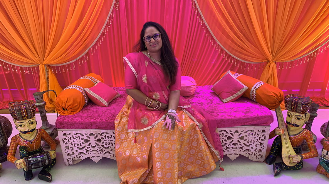 Reena is an Indian woman, wearing a sari. She is sitting on pink cushion on a white bench, in a room with bright pink and orange draping. She is wearing glasses and smiling with a closed mouth. 