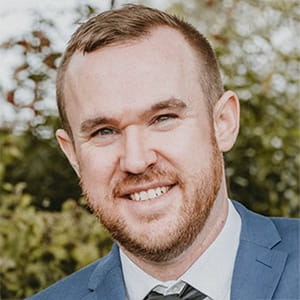 Ryan Kelly has auburn hair and is wearing a light blue suit jacket, blue tie and white shirt. There are trees in the background.