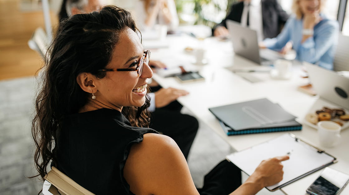 Over the shoulder profile of bespectacled female executive in early 30s sitting at conference table and laughing as she interacts with off-camera colleague.