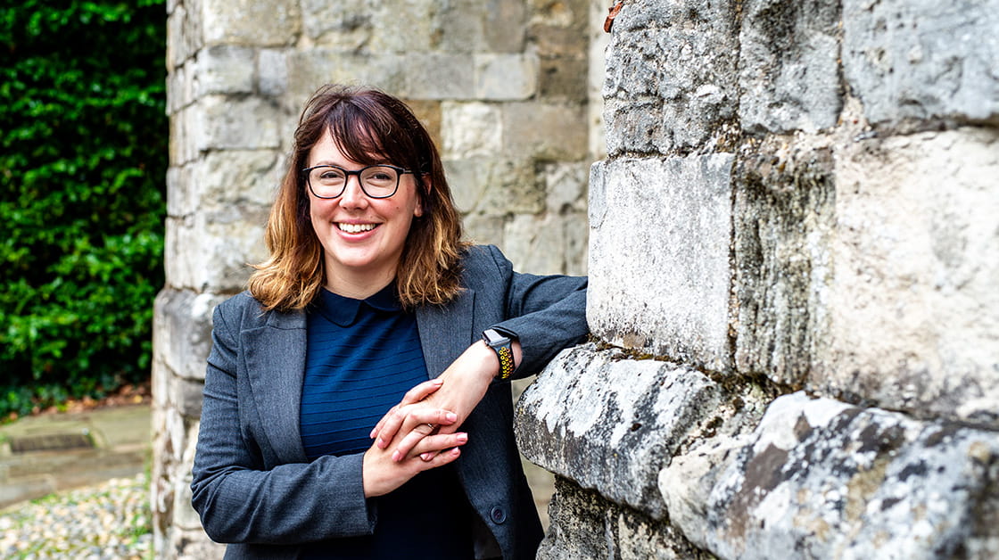 Chloe Jay is a white woman smiling and leaning an elbow on a weathered stone building. She wears glasses, a grey blazer and dark top and has shoulder-length brown hair.