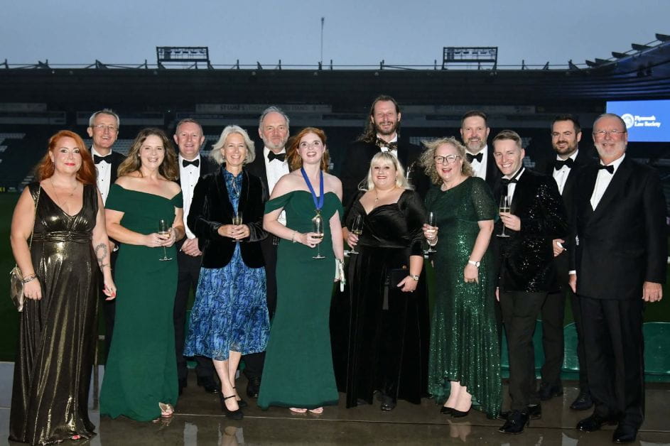 A large group of solicitors stand together smiling. They are at a black tie event with the men dressed in tuxedos in the women in long formal dresses.