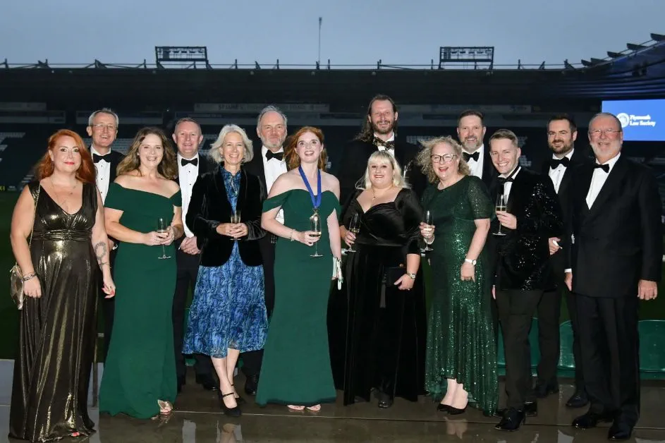 A large group of solicitors stand together smiling. They are at a black tie event with the men dressed in tuxedos in the women in long formal dresses.