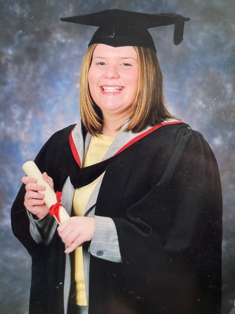 Rebecca Wood smiles while wearing graduation robes and mortar board. She is holding a degree scroll.