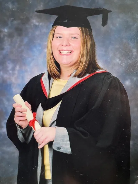 Rebecca Wood smiles while wearing graduation robes and mortar board. She is holding a degree scroll.