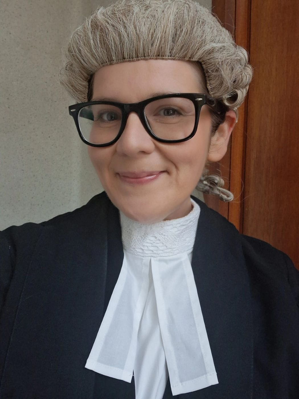 Rebecca smiles whilst wearing formal court attire including a wig. She also wears glasses with a dark frame.