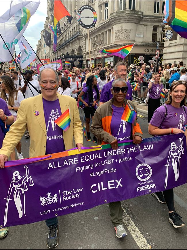 A white man, black woman and white woman are walking and smiling, holding a blue banner. They are outside in front of a crowd.