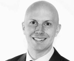 Chris is a white man with no hair. The photo is in black and white, and he stands in front of a white background wearing a suit and tie. He is smiling.