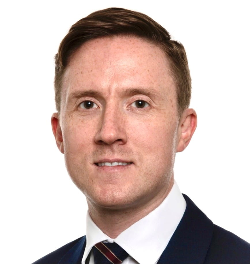 Eoghan (pronounced oh when) is a white man with light brown hair. He is wearing a suit with a white shirt and tie, and stands in front of a plain white background.