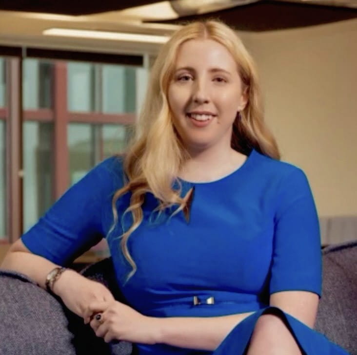 Georgia is a white woman with blonde hair. She is wearing a blue dress and is smiling.