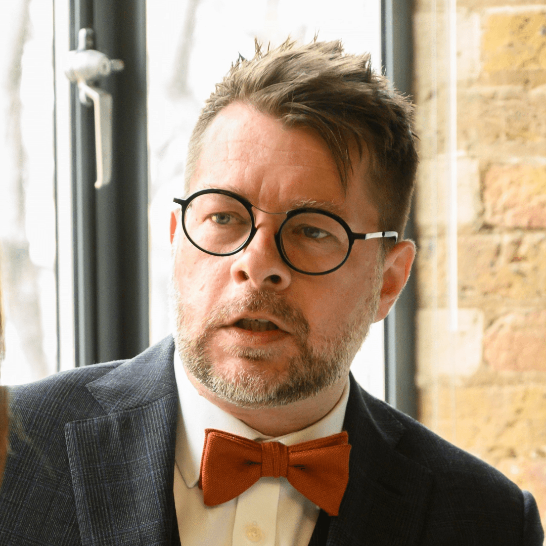 Johnathan is a white man. He is wearing a suit with a red bow tie and he is in front of a window. 