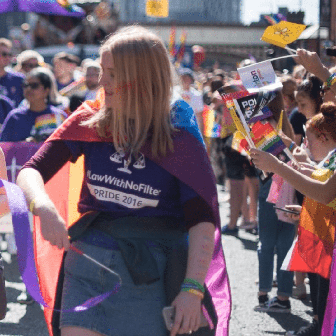 Laura is a white woman. She is wearing a purple t-shirt with a Pride flag on her back. She is standing in a crowd of people. 