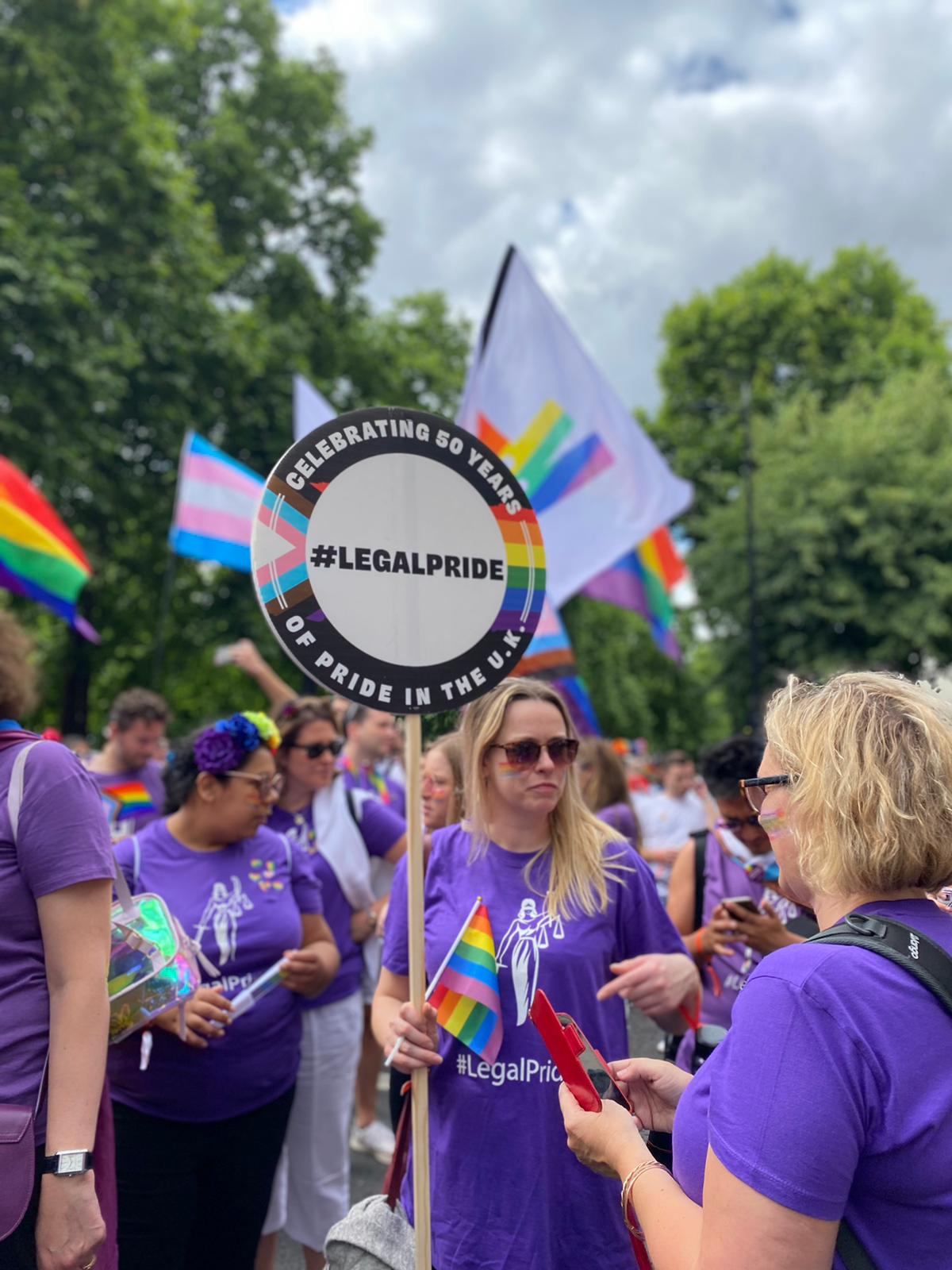 A blonde, white woman is wearing sunglasses and standing amongst a group of people. She is holding a sign which says hashtag Legal Pride. 