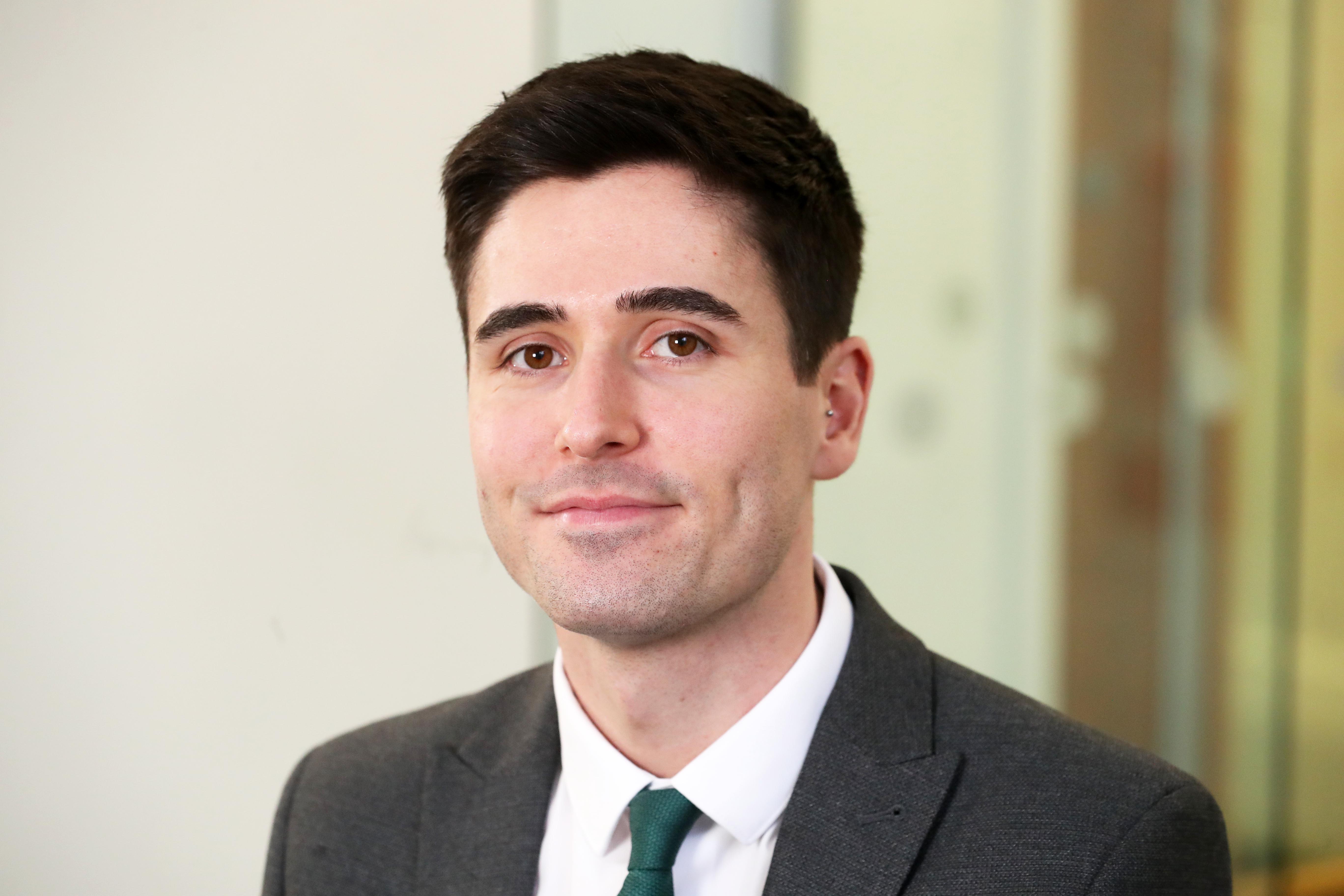 Sam is a white man with dark hair. He is wearing a grey suit with a white shirt and a green tie, standing in front of a plan background.