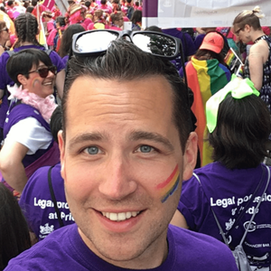 Tom is a white man with a rainbow painted on his face. He is smiling in front of a crowd of people.