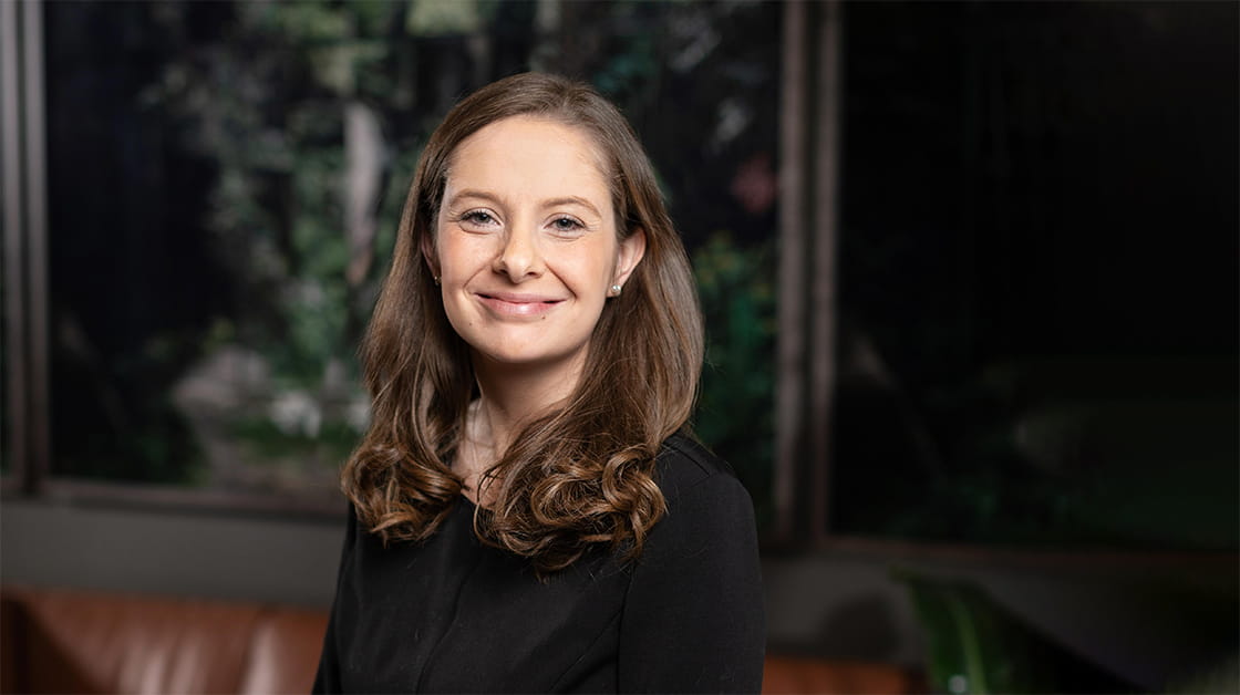 Laura Uberoi stands smiling in a dark room in front of a framed landscape. She is a white woman with long, wavy brown hair, wearing a black blouse, black-and-white polka dot skirt and pearl earrings.