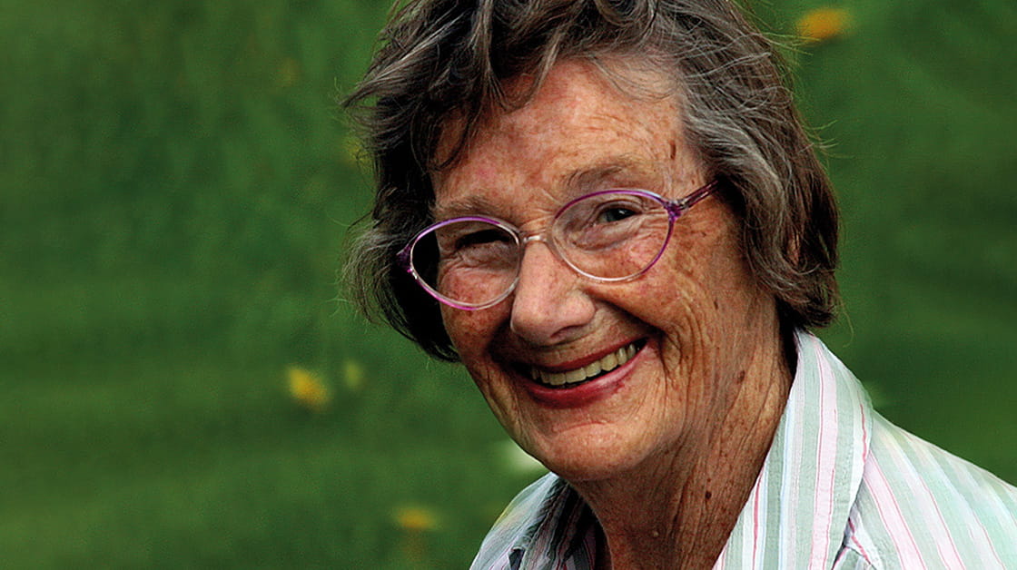Joan Blass was a white woman with short, wavy dark grey hair. She is smiling and wears glasses and a sage green, white and pink striped shirt.