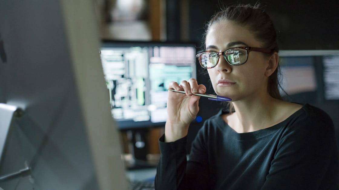 A woman looks at a computer screen, the screen reflects on the lenses of her glasses.