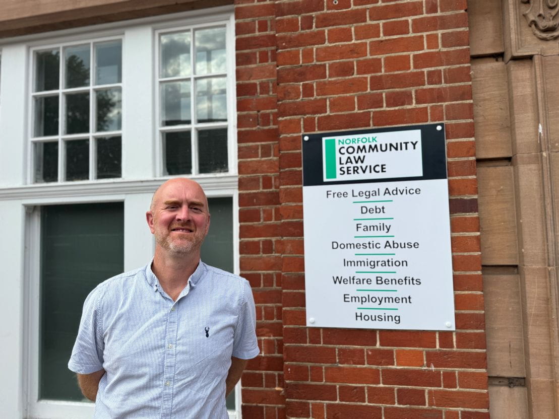 Man standing in front of a brick building with a sign for Norfolk Community Law Service listing free legal services