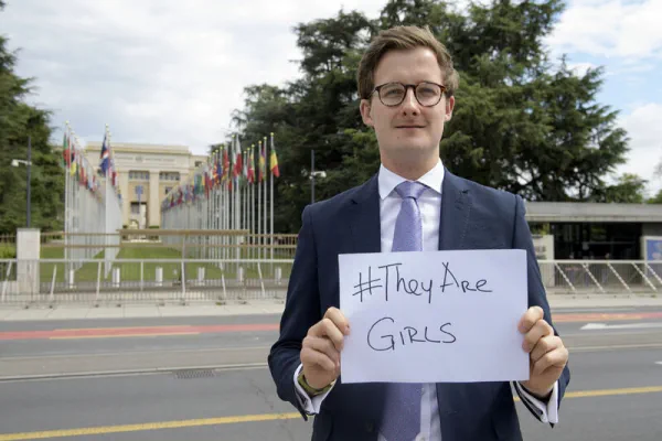 A white man with brown hair and glasses, wearing a blue suit, stands outside the UN holding a sign saying hashtag they are girls.