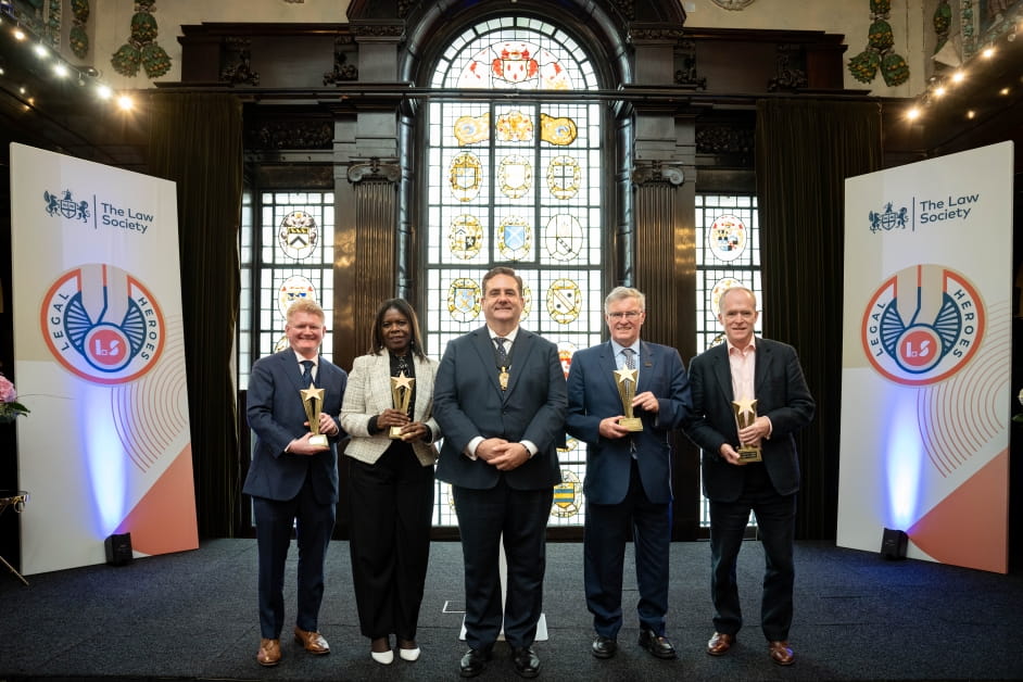 Five smartly dressed people stand on stage at the Legal Heroes awards ceremony. Four of the people are holding awards.