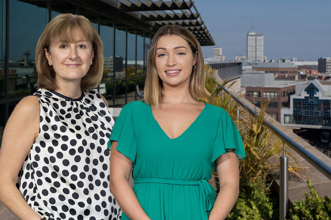 Susan Howe and Georgia Barber, standing on balcony of Muckle LLP's office in Newcastle on a sunny day. Both are white women with shoulder-length hair, Susan is older and wears a sleeveless white top with black polkadot and Georgia is in her early 20s and wears a mint-green jumpsuit. Behind them is a glass building, decking and outdoor plants with Newcastle buildings in the background.