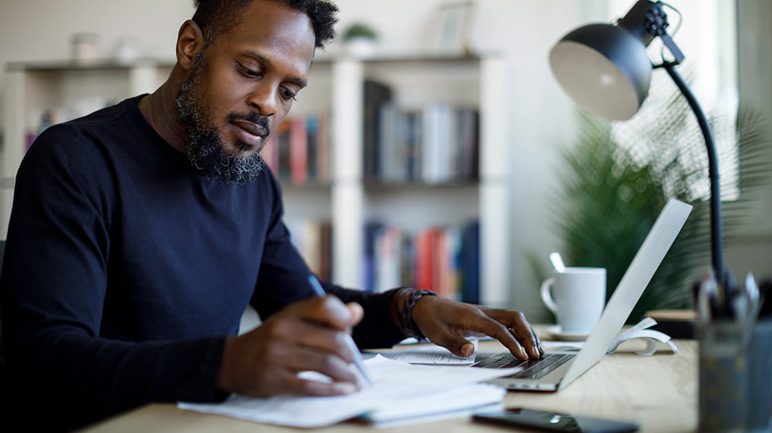 A Black professional man with short, black afro hair and a beard sits at a desk writing in a notebook with a pen. He is concentrating and wears a navy jumper.