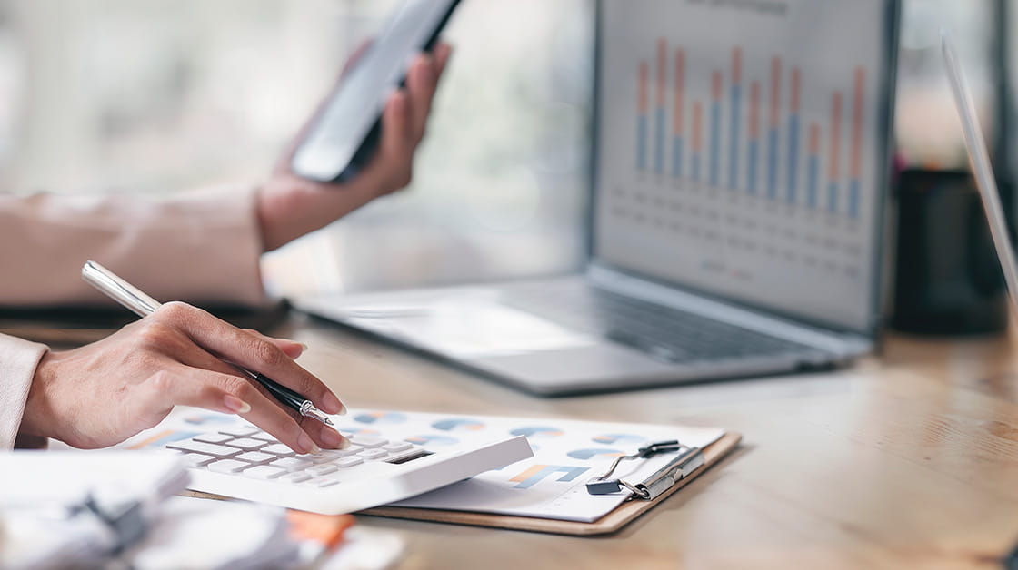 A person manages their finances using a calculator and financial projections on a laptop.