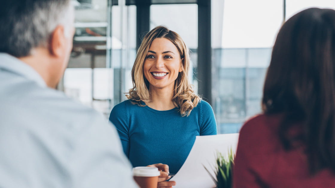A white woman with medium length blonde hair is sitting opposite a couple and smiling. She is wearing a blue top and holding some papers. 
