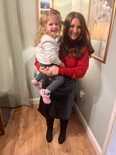 Solicitor Emma Chater holds her daughter up on her hip and they both smile. Emma wears a red top and black skirt whilst he daughter wears a light coloured top