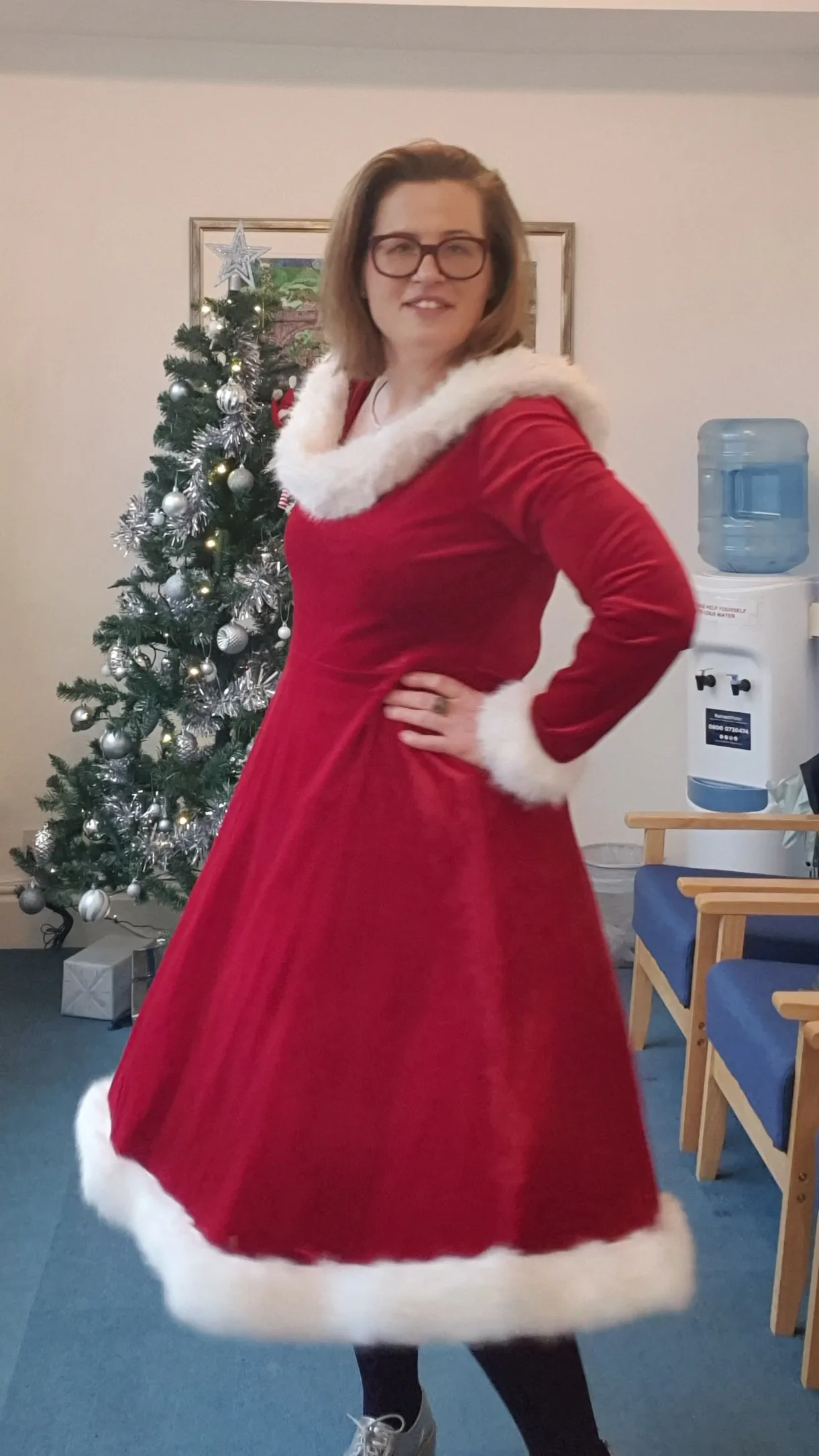 A woman dressed as Mrs Claus stands in front of a Christmas tree.