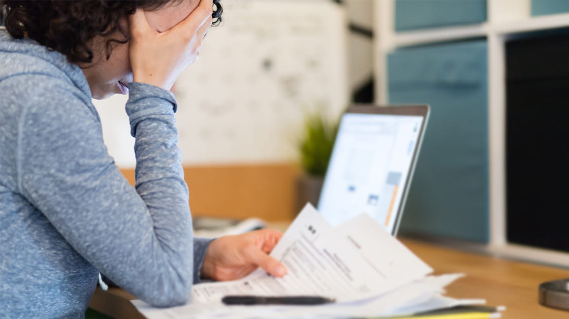 A white woman with dark, curly hair wearing a blue top sits at a desk looking at a bill, with a calculator open on her laptop. She has her head in her hands and looks stressed by her finances.