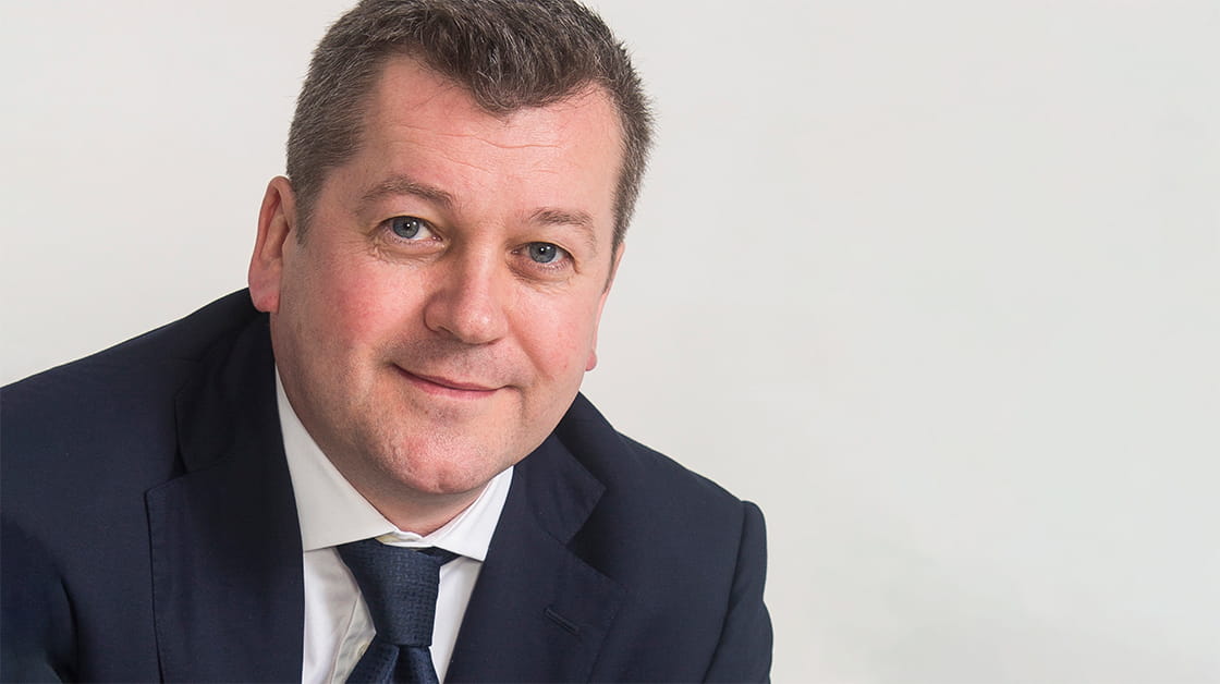 Jon Goodwin, a white man with short brown hair, sits smiling and wearing a navy suit, white shirt and navy tie.
