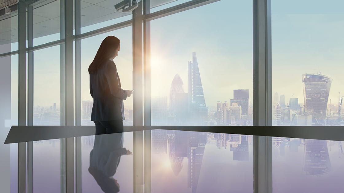 Silhouette of woman holding phone looking out modern office window at City of London skyline at early morning. She has long straight hair and is wearing a loose blazer.