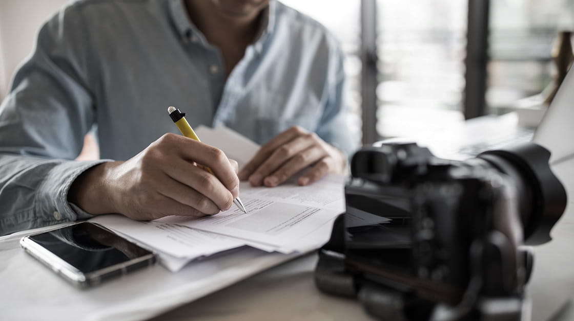 A man with a camera on his desk in the foreground signs a contracts.