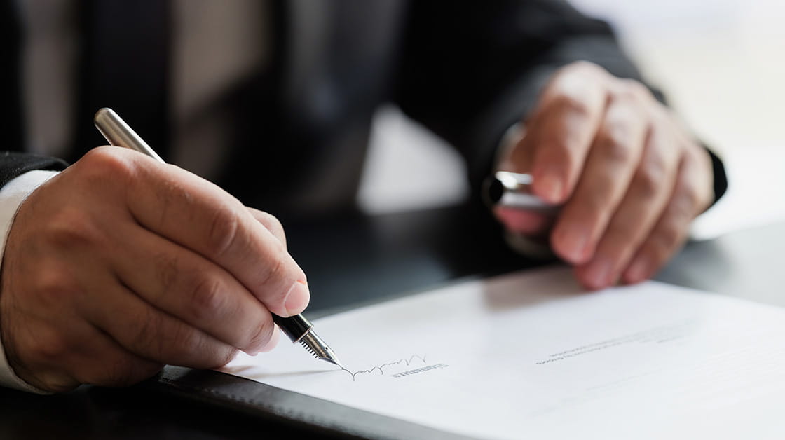 A person signs a hard-copy document using a fountain pen. They wear a dark suit, white shirt and dark tie.